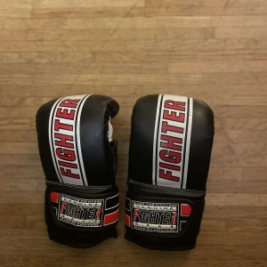 boxningshandskar Fighter - Säljer ett par svarta boxningshandskar från Fighter med vita och röda detaljer. Handskarna har en robust design med justerbara kardborreband för optimal passform. Perfekta för träning och sparring.