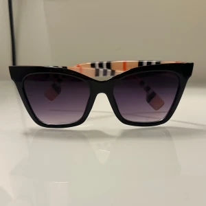 Svarta solglasögon från Burberry - Snygga svarta solglasögon från Burberry med en klassisk fyrkantig form. Bågarna har en stilren design med Burberrys ikoniska rutmönster på insidan av skalmarna i svart, vitt och orange. Perfekta för att ge en touch av lyx till din stil.