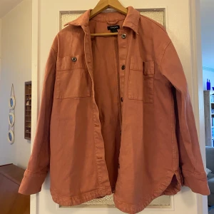 Rostfärgad overshirt från Lindex - Snygg rostfärgad overshirt från Lindex i en avslappnad stil. Skjortan har långa ärmar och knappar framtill samt två bröstfickor med knappdetaljer. Perfekt för lager-på-lager-look!