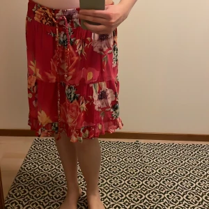 Blommig kjol med volang - Säljer en färgglad blommig kjol i rosa med volangdetaljer. Kjolen har ett knytband i midjan och är perfekt för sommarens varma dagar. Den har en lös passform och är gjord i ett lätt material.
