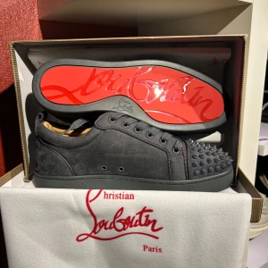 Grå skor från Christian Louboutin - Snygga grå sneakers i mocka från Christian Louboutin med den ikoniska röda sulan. Skorna har snörning och är dekorerade med nitar på tån för en edgy look. Perfekta för dig som vill sticka ut med stil.OBS! Priset är inte hugget i sten. Hör av er vid minsta fundering 🍾