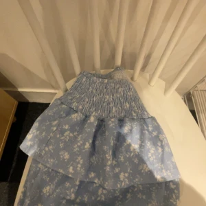 Blå blommig kjol - Charmig blå kjol med blommigt mönster och volangdetaljer. Perfekt för en somrig look med sin lätta och luftiga design.