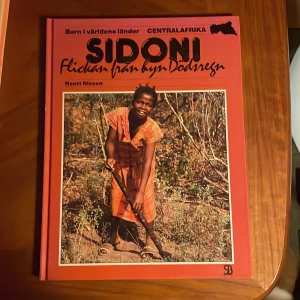 Sidoni: Flickan från byn Dödsgregn - Boken 'Sidoni: Flickan från byn Dödsgregn' av Henri Nissen är en del av serien 'Barn i världens länder' och fokuserar på Centralafrika. Den har en röd framsida med en bild av en leende flicka i en orange klänning. Boken utforskar livet i Centralafrikanska republiken.