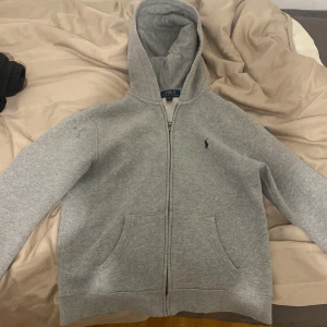 Grå hoodie från Polo Ralph Lauren - Storlek l i barnstorlek-16-18. Säljer en klassisk grå hoodie från Polo Ralph Lauren med dragkedja och huva. Tröjan har långa ärmar och en liten broderad logga på bröstet. Perfekt för en avslappnad stil. 🐎