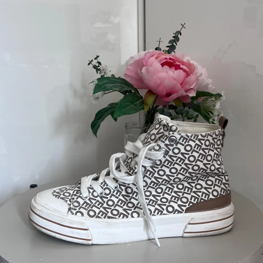 Säljer ett par coola vita sneakers från Graceland med ett unikt textmönster där det står 'LOVE' över hela skon. De har en brun detalj runt hälen och en vit sula med bruna ränder. Skorna har snörning framtill och är så gott som nyskick. Perfekta för vår och sommar!. Skor.