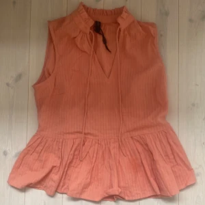 Persikofärgad ärmlös topp från YAS - Säljer en superfin persikofärgad topp från YAS. Den är ärmlös och har en söt volangdetalj längst ner som ger en härlig sommarkänsla. Toppen har en v-ringning med knytband.står inte vad de är för storlek men skulle tror att de är s.använd 1 gång