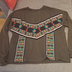 Brun tröja med färgglatt mönster - Säljer en unik armgreen  tröja med ett färgglatt geometriskt mönster i rött, turkos och orange. Tröjan har långa ärmar och en normal passform. Perfekt för att sticka ut i mängden!