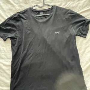 Svart t-shirt från Hugo Boss - Säljer en stilren svart t-shirt från Hugo Boss. Den är i bra skick och har en klassisk design med korta ärmar och rund hals. Perfekt för både vardag och fest!
