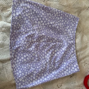 Lila blommig kjol - Superfin lila kjol med ett sött blommönster i vitt. Perfekt för sommardagar eller en vårutflykt. Kjolen är kort och har en normal passform, vilket gör den både bekväm och stilren. Ett måste i garderoben för alla som älskar blommiga mönster!