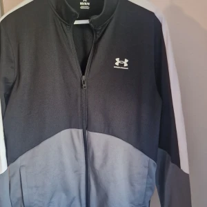 Svart och grå jacka från Under Armour - Säljer en snygg svart och grå jacka från Under Armour. Den har en dragkedja framtill och vita detaljer på ärmarna. Perfekt för träning eller vardagsbruk! Används bara en gång säljar pga vikt.