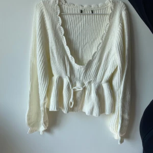 Vit stickad kofta med volangdetaljer - Säljer en superfin vit stickad kofta med volangdetaljer längs med den v-ringade halsen. Den har långa ärmar och ett knytband i midjan för en snygg passform. Perfekt för både höst och vår när du vill ha något mysigt men ändå stilrent. Passar till både jeans och kjol!