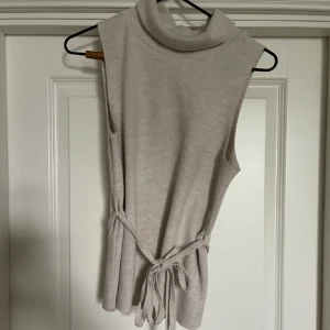 Beige ärmlös polotopp med knytband - Säljer en stilren beige ärmlös tröja med krage med ett snyggt knytband i midjan från zara. Perfekt för både vardag och festliga tillfällen. Toppen är i ett mjukt och bekvämt material som ger en elegant look.