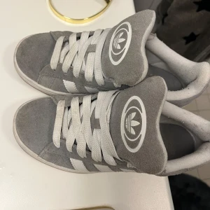 Grå sneakers från Adidas - Säljer ett par snygga grå sneakers från Adidas. De har en klassisk design med vita ränder på sidorna och det ikoniska Adidas-loggan på tungan och hälen. Skorna är i mocka och har en bekväm passform med snörning. Perfekta för vardagsbruk!
