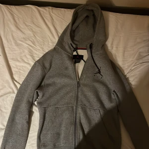 Grå hoodie från Jordan - Säljer en snygg och bekväm grå hoodie från Jordan. Den har en dragkedja framtill och en mjuk insida som håller dig varm. Perfekt för både vardag och träning. Hoodien har långa ärmar och en klassisk passform. Passa på att köpa den här stilrena tröjan!