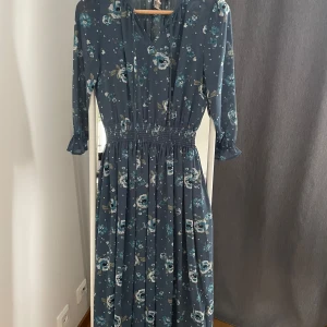 Blommig långklänning från H&M Divided - Säljer en superfin långklänning från H&M Divided i storlek EU 32. Klänningen är blå med ett vackert blommigt mönster i olika nyanser av blått och vitt. Den har trekvartsärmar och en smockad midja som ger en fin passform. Perfekt för vår och sommar!