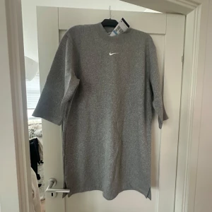 Grå klänning från Nike - Säljer en stilren grå klänning från Nike. Den har en enkel design med en liten vit Nike-logga på bröstet. Klänningen är i ett mjukt material och har trekvartsärmar. Perfekt för en avslappnad stil eller träning. Den har en liten slits på sidan för extra rörelsefrihet.