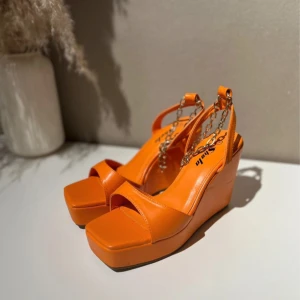 Orange kilklackssandaler med kedjedetalj - Nya! Säljer ett par snygga orange sandaler med kilklack och coola kedjedetaljer. Perfekta för sommaren och att liva upp vilken outfit som helst! De har en öppen tå och är i syntetmaterial. 