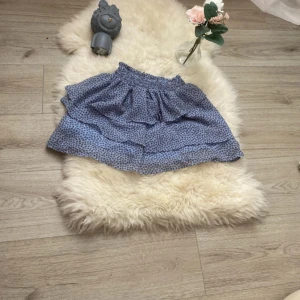 Blå blommig kjol från H&M - Säljer en superfin blå kjol från H&M med ett sött blommigt mönster. Kjolen har en volangdetalj och är perfekt för sommarens alla äventyr. Den är i storlek 9-10 år och har en bekväm resår i midjan. Passar perfekt till både vardag och fest!