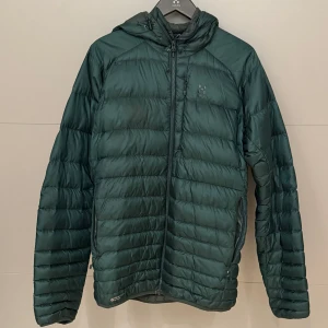 Grön dunjacka från Haglöfs - Säljer en snygg och varm grön dunjacka från Haglöfs. Den har en praktisk huva och dragkedja framtill. Perfekt för kyliga höst- och vinterdagar. Jackan är i bra skick och redo för nya äventyr!