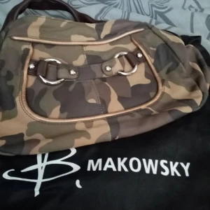 Camouflageväska från B. Makowsky - Snygg axelväska i camouflage från B. Makowsky. Väskan har ett coolt mönster och är perfekt för en avslappnad stil. Den har en dragkedja och detaljer i brunt skinn. Insidan är fodrad med leopardmönster, vilket ger en extra touch av stil. Perfekt för vardagsbruk!