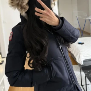 Mörkblå dunjacka med pälsdetaljer - Säljer en snygg mörkblå dunjacka med pälsdetaljer på huvan. Jackan har dragkedja och praktiska fickor med knappar. Perfekt för kalla vinterdagar och håller dig varm och stilren. Passar både till vardags och lite finare tillfällen.