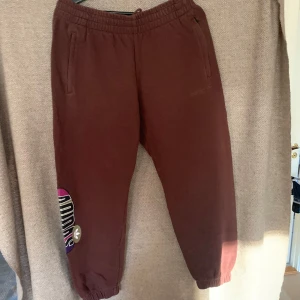 Bruna mjukisbyxor från Adidas - Säljer ett par bruna mjukisbyxor från Adidas i storlek L. De har en elastisk midja och muddar vid bensluten för en bekväm passform. Byxorna har en cool logga på sidan i färgglada nyanser. Perfekta för en avslappnad stil eller träning!