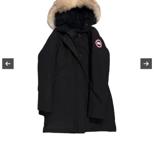 Svart parkajacka från Canada Goose/ victoria parka  - Säljer en svart parkajacka från Canada Goose i mycket bra skick. Jackan har en varm pälskantad huva och är perfekt för kalla vinterdagar. Den har en klassisk design med lång ärm och en snygg logga på ärmen. Perfekt för både stad och natur!