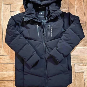 Svart dunjacka från Sail Racing - Säljer en svart dunjacka från Sail Racing i mycket bra skick. Jackan har en stilren design med dragkedja och flera praktiska fickor. Perfekt för kalla vinterdagar med sitt hypoallergena dunfoder. Den är tillverkad enligt OEKO-TEX standard och har en fyllkraft på 500. Passar både till vardags och utomhusaktiviteter.