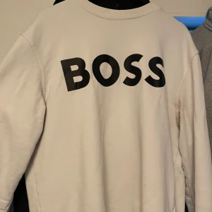 Vit sweatshirt från BOSS - Säljer en stilren vit sweatshirt från BOSS i mycket bra skick. Den har en stor BOSS-logga på framsidan och är perfekt för både vardag och avslappnade tillfällen. Tröjan är långärmad och gjord i ett mjukt material som känns skönt mot huden. Passar perfekt till jeans eller joggers!