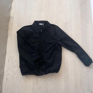 Svart jacka med dragkedja - Säljer en stilren svart jacka från Weekday i mycket bra skick. Jackan har en dragkedja framtill och två bröstfickor med lock. Perfekt för höst och vår, och passar till de flesta outfits. Den är långärmad och har en normal passform. 