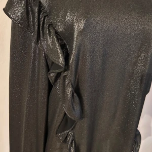 Svart glittrig blus från KappAhl - Snygg svart blus från KappAhl i storlek 40. Den har en glittrig yta och volangdetaljer längs sidorna och nederkanten. Perfekt för festliga tillfällen med sina långa ärmar och eleganta stil. Blusen är i mycket bra skick och redo att bäras på nästa party! ✨
