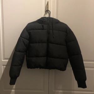 Svart pufferjacka från Monki - Säljer en helt oanvänd pufferjacka från Monki, nypris närmare 600kr. Storlek xxs men passar även xs! Den är kortare i modellen och har en mysig huva för extra värme. Perfekt för både höst och vinter!