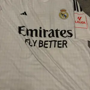 Säljer en vit Real Madrid fotbollströja från Adidas i mycket bra skick. Tröjan har korta ärmar och är dekorerad med klubbens emblem och sponsorlogga. På baksidan finns namnet 'VINI JR.' och nummer 7. Perfekt för alla fans av laget!