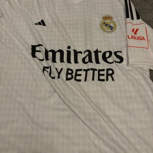 Vit Real Madrid fotbollströja - Säljer en vit Real Madrid fotbollströja från Adidas i mycket bra skick. Tröjan har korta ärmar och är dekorerad med klubbens emblem och sponsorlogga. På baksidan finns namnet 'VINI JR.' och nummer 7. Perfekt för alla fans av laget!
