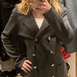 Grå kappa från Joy of Love - Säljer en snygg grå kappa från Joy of Love i storlek S. Den har en klassisk design med dubbelknäppning och stora knappar. Perfekt för höst och vår, och ger en stilren look. Kappan är i mycket bra skick och passar både till vardags och lite finare tillfällen.