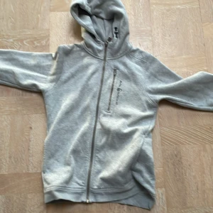 Grå hoodie från Sail Racing - Säljer en snygg grå hoodie från Sail Racing i bra skick. Den har en dragkedja framtill och en praktisk ficka med dragkedja på bröstet. Perfekt för höst och vår, med en bekväm passform och huva för extra mysfaktor. Perfekt för både vardag och träning! Ny pris 1400kr (Ej använd mycket)