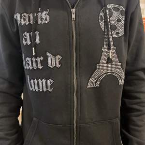 Säljer en svart hoodie med dragkedja och coolt Paris-motiv i glittriga stenar. Perfekt för dig som vill ha en unik stil! Den har långa ärmar och en bekväm passform. Passar både till vardags och festliga tillfällen. 🖤