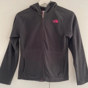 Svart fleecejacka från The North Face - Säljer en svart fleecejacka från The North Face i mycket bra skick. Den har en dragkedja i rosa och en huva för extra mys. Perfekt för höst och vår när det är lite kyligt ute. Fleecen har långa ärmar och är supermjuk och bekväm. Passar perfekt för både vardag och utomhusaktiviteter!