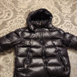 Svart dunjacka från Moncler - Säljer en snygg svart dunjacka från Moncler i mycket bra skick. Jackan har en glansig finish och är perfekt för kalla vinterdagar. Den har en huva och dragkedja framtill, samt praktiska fickor med dragkedjor. Insidan har ett coolt serietidningsmönster. Perfekt för dig som vill hålla stilen i vinter! 🖤