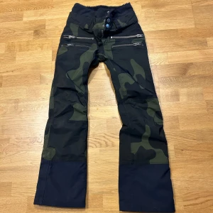 Camouflage byxor från SOS Black Snow - Säljer ett par coola camouflagebyxor från SOS Black Snow i storlek 36 S. Byxorna har en grön och svart camo-look med dragkedjor och knappar framtill. Perfekta för vintern med sin robusta design och höga midja. Passar både för vardag och utomhusaktiviteter.