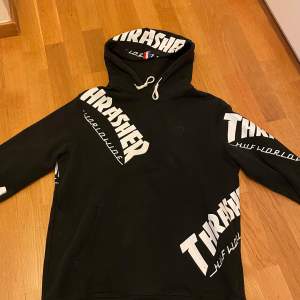 Säljer en svart hoodie från Thrasher med vit text och broderad logga. Den har en stor huva med dragsko och är perfekt för en avslappnad stil. Passar bra till både vardag och skate. Storleken är inte angiven, men den ser ut att ha en normal passform.