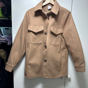 Beige overshirt med knappar - Säljer en snygg beige overshirt i mycket bra skick. Den har en klassisk krage och stora knappar framtill. Två bröstfickor med lock ger en cool look. Perfekt för höst och vår, och funkar både till vardags och lite finare tillfällen. Passar bra över en t-shirt eller tröja.