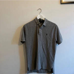 Grå pikétröja från Ralph Lauren - Säljer en klassisk grå pikétröja från Ralph Lauren. Den har en broderad logga på bröstet och knappar vid kragen. Perfekt för en stilren och avslappnad look. Passar både till vardags och lite finare tillfällen.