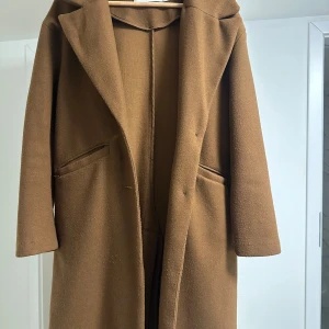 Brun kappa - Säljer en stilren brun kappa i ull. Perfekt för höst och vinter med sin klassiska design och långa ärmar. Den har en snygg krage och två fickor framtill. Passar både till vardags och festliga tillfällen. Ett måste i garderoben för den som vill hålla sig varm och trendig!