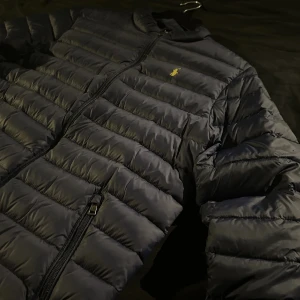 Svart dunjacka från Ralph Lauren - Säljer en snygg svart dunjacka från Ralph Lauren. Jackan har en dragkedja framtill och är perfekt för kyliga dagar. Den är lätt och bekväm med en liten gul logga på bröstet. Passar både till vardags och lite finare tillfällen. Perfekt för höst och vinter. Storleken är 170/xxs
