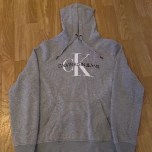 Grå hoodie från Calvin Klein Jeans - Säljer en snygg grå hoodie från Calvin Klein Jeans i storlek M. Den har en klassisk design med stor logga på framsidan och en bekväm huva med dragsko. Perfekt för en avslappnad stil och passar till alla tillfällen. Skön och stilren!