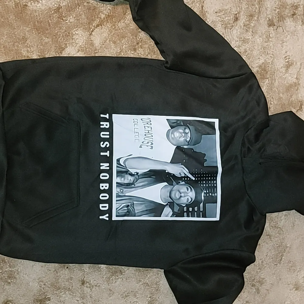 Säljer en svart hoodie med coolt tryck på framsidan och texten 'Trust Nobody'. Baksidan har en lista med Tupac Shakur-låtar. Perfekt för dig som gillar streetwear och vill ha något unikt i garderoben. Den är i bra skick och superbekväm att ha på sig. Passar perfekt till höst och vinter!. Tröjor & Koftor.