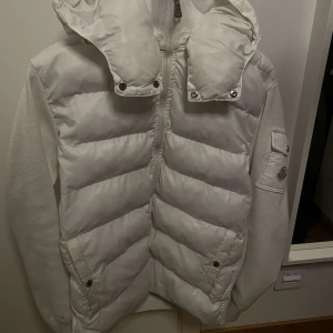 Vit pufferjacka från Moncler - Säljer en snygg vit pufferjacka från Moncler. Jackan har en quiltad design med dragkedja och en cool broderad logga på ärmen. med sin varma och stiliga look. Den har också en praktisk innerficka med en rolig serieteckning. Passar perfekt för både vardag och fest!      Obs! liten hål i insidan av luvan som inte syns ut kan skicka gärna bild på den😃