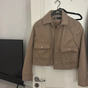 Beige kappa från Zara - Säljer en snygg beige kappa från Zara i mycket bra skick. Den har en klassisk krage och långa ärmar. Jackan är croppad och har två stora fickor framtill med knappar. Perfekt för höst och vår!