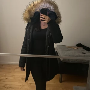 Svart parkajacka med pälsfodrad huva - Säljer en svart parkajacka från SMG4 i mycket bra skick. Jackan har en mysig pälsfodrad huva och stängs med dragkedja och knappar. Perfekt för kalla vinterdagar! Den har en amerikansk flagga som detalj på ärmen. Jackan är i strl Large men funkar fint för mig som bär S-M